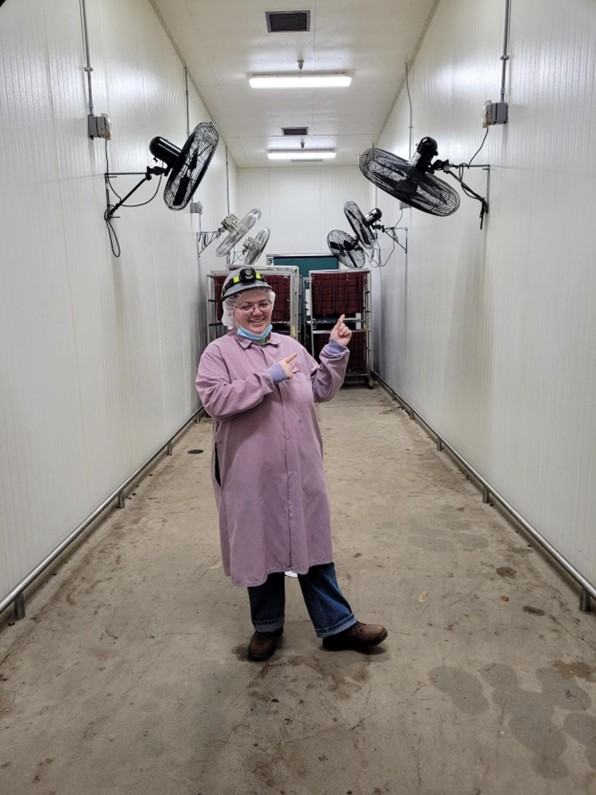 Hallie stands in a room with multiple suspended fans. She wears a lab coat, a hair net, and a hard hat.