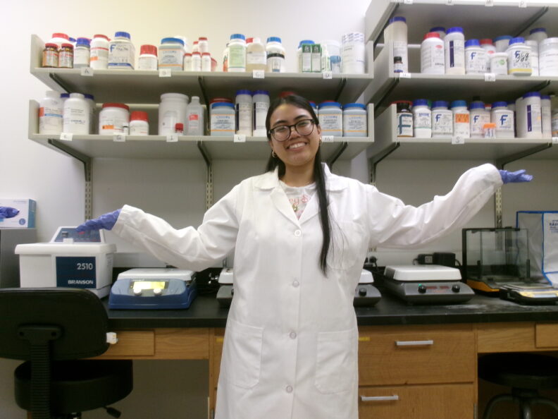 Sherine in a lab coat standing in front of shelves of bottled chemicals.
