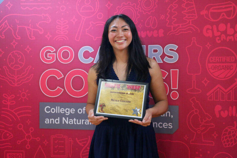 Natalie holds up her award and smiles, standing in front of a red background.