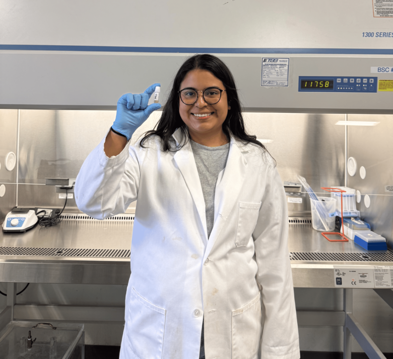 Miriam wears a lab coat and gloves, holding up a vial.