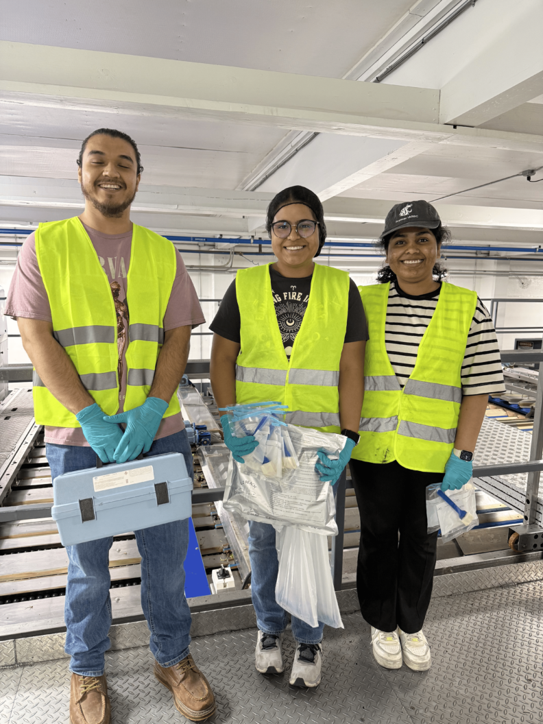 Miriam and two other students wear high-vis vests. They all hold different things. Miriam holds a plastic bag full of paper, the student on her left holds a toolbox, and the student on her right holds a plastic bag with a brush inside.