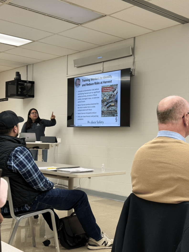 Miriam stands at a podium and presents a slide titled, "Training Workers to Identify and Reduce Risks at Harvest."