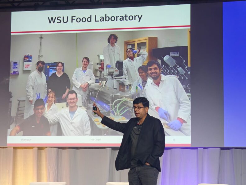 Dr. Ganjyal motions to his presentation that shows a slide of his lab group. Two students give Dr. Ganjyal bunny ears and another poses dramatically against the extrusion machine.