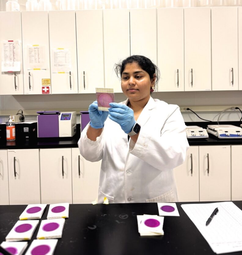 Swapnika stands in a lab and holds up a petrifilm plate.