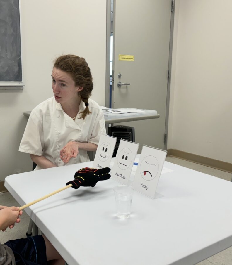 Rachel talking to a child. The child uses a toy hand on a stick to point to three different signs. The signs say "yummy, just okay, and yucky."