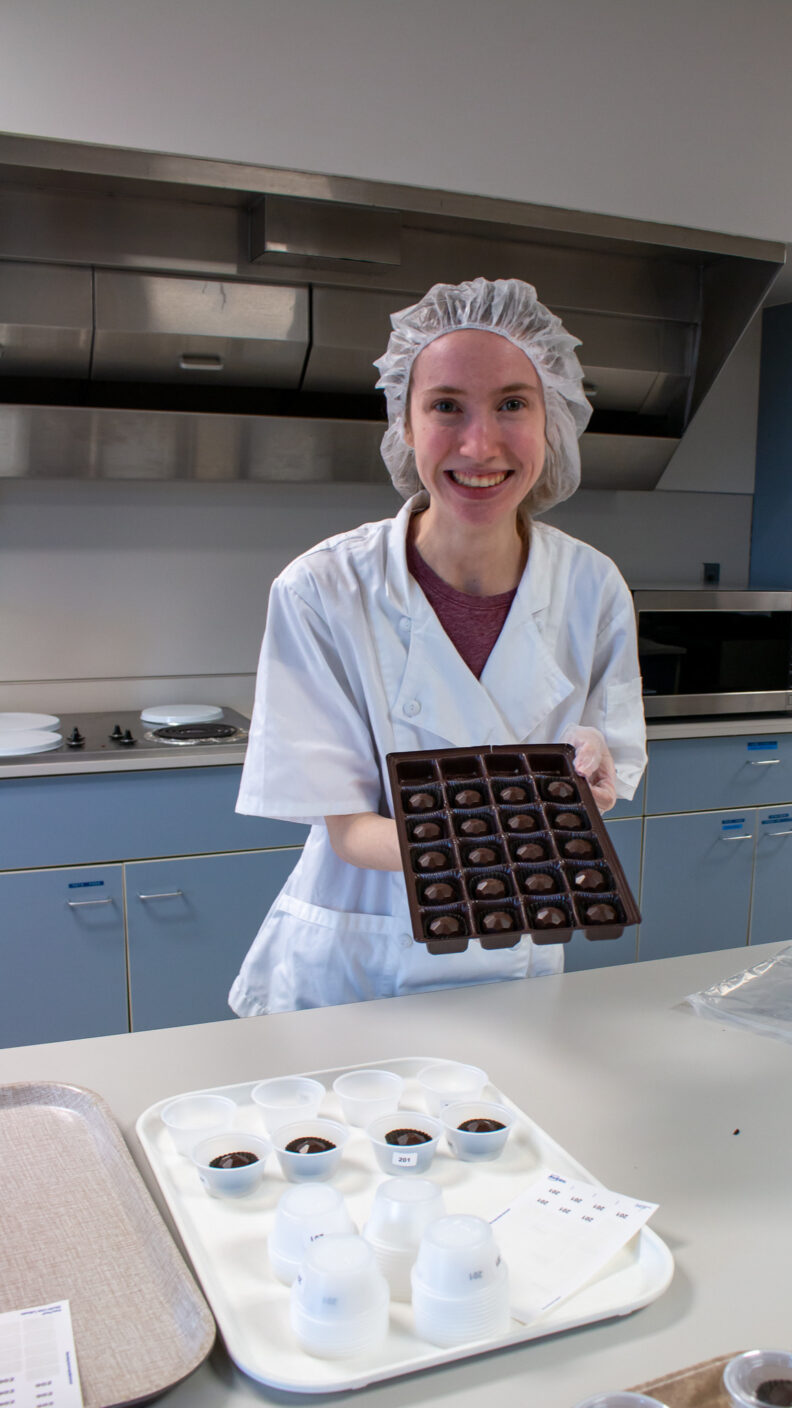 Rachel holds a tray full of chocolates. She wears a lab coat and a hair net.