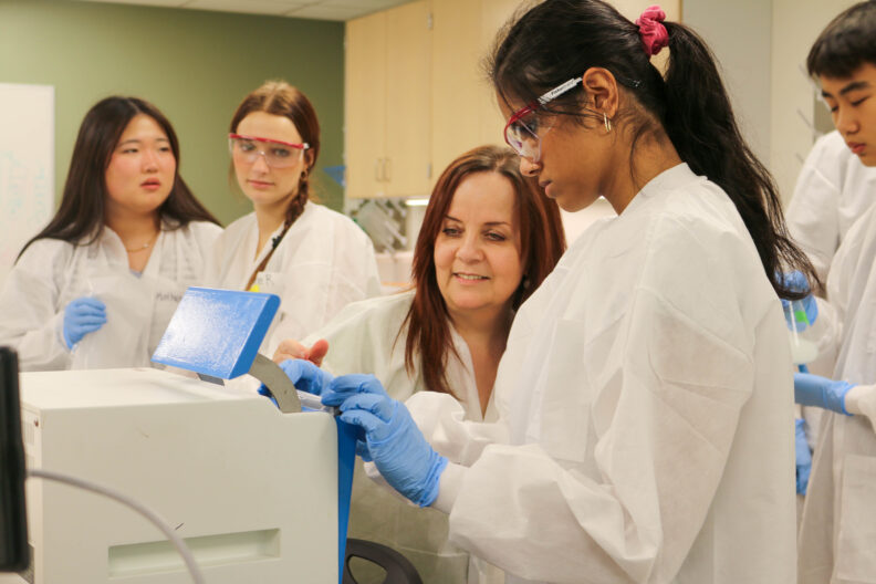 Gulhan and a student stand by a white machine. Other students linger around them. Everyone wears a lab coat.