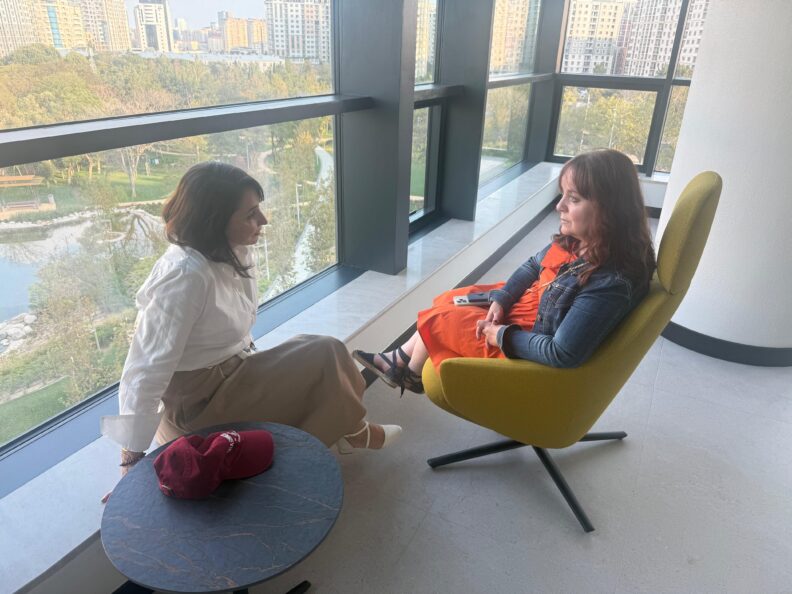 Two women sit beside each other in discussion. One sits in a chair and the other sits on a ledge.