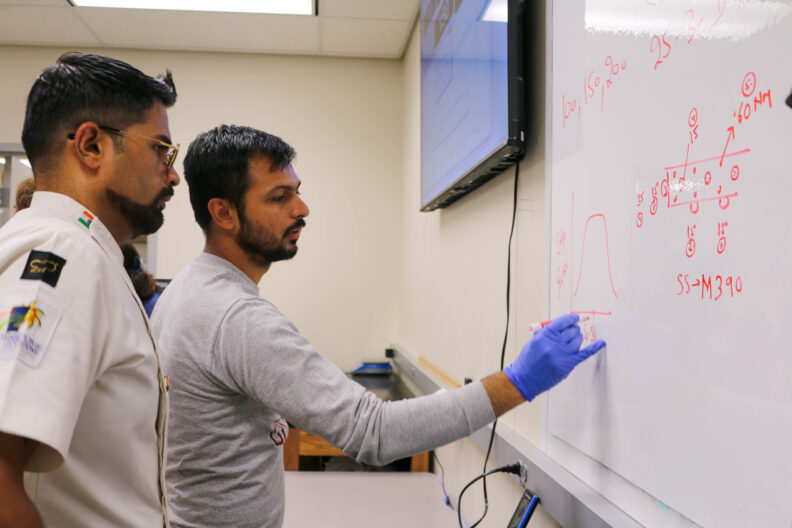 Aniket writes on a whiteboard in market. Other notes are written on the board. He labels a bell curve. A man stands beside Aniket, listening to what he has to say.