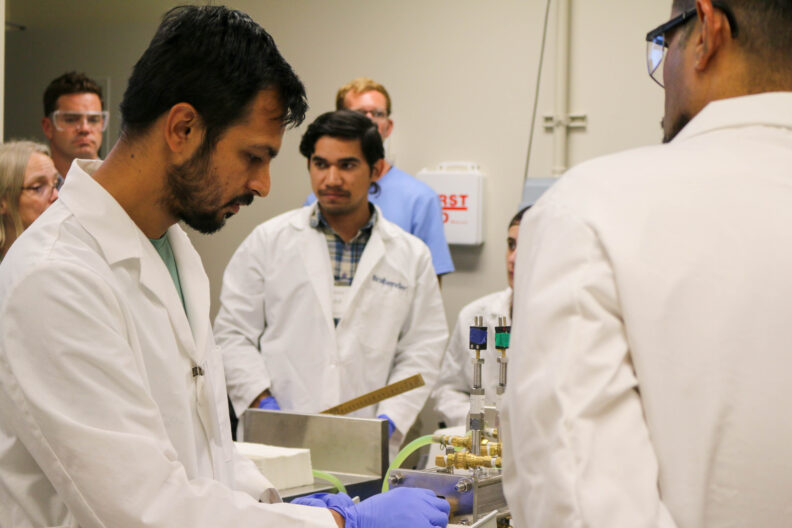 Aniket looks down at the end of the extrusion machine as it processes material. A group of people are in the room.
