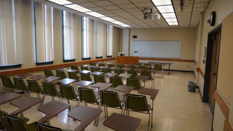 Looking out from the rear of the classroom over the rows of student seating and toward the front of the room where the instructor's podium and one whiteboard are located.