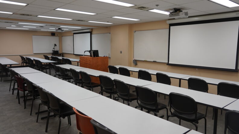 Looking out from the rear corner of the classroom over the rows of student seating and toward the front of the room where the instructor's podium, two display screens, and two wall-mounted whiteboards are located.