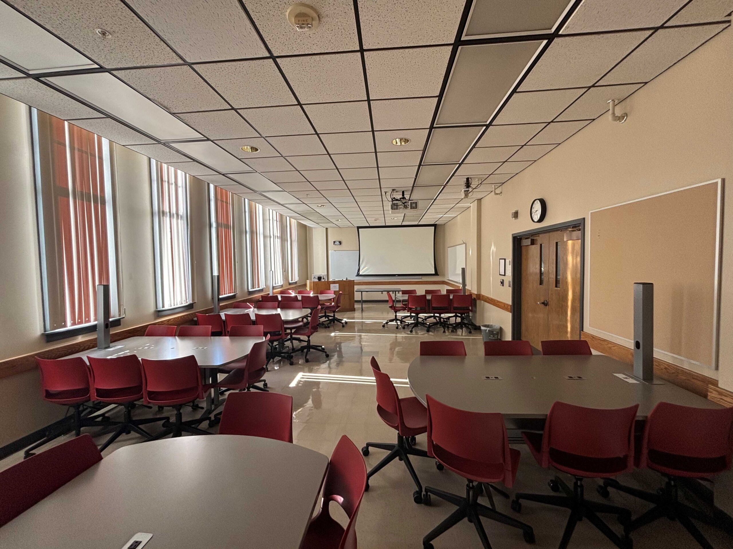 View from the back of the classroom showing six D‑shaped tables, each with six chairs. At the front of the room is an instructor podium with technology next to a projection screen. Double doors leading out of the classroom are centered on the right wall.