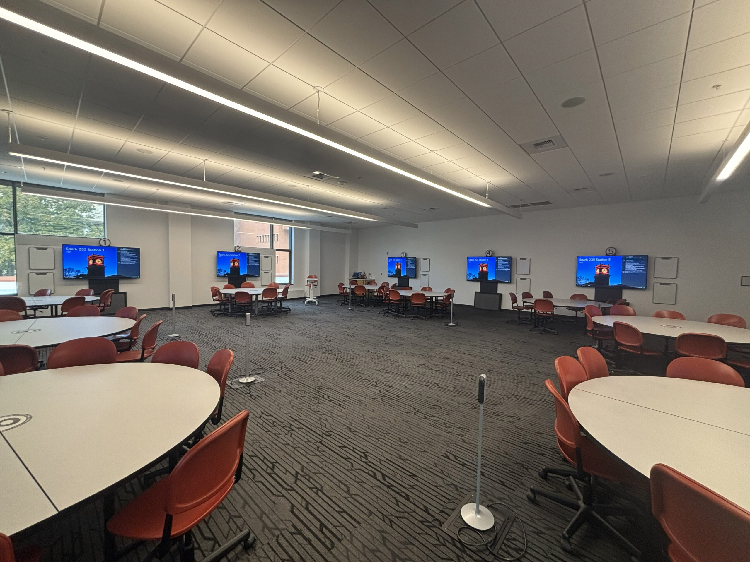 Looking out from the corner of the classroom over the mobile instructor lectern and nine student groups which consist of a round table with nine chairs and a large monitor for viewing content. There are several small rectangular mobile whiteboards hanging on the walls as well as five convenience power towers available for plugging in devices around the room.