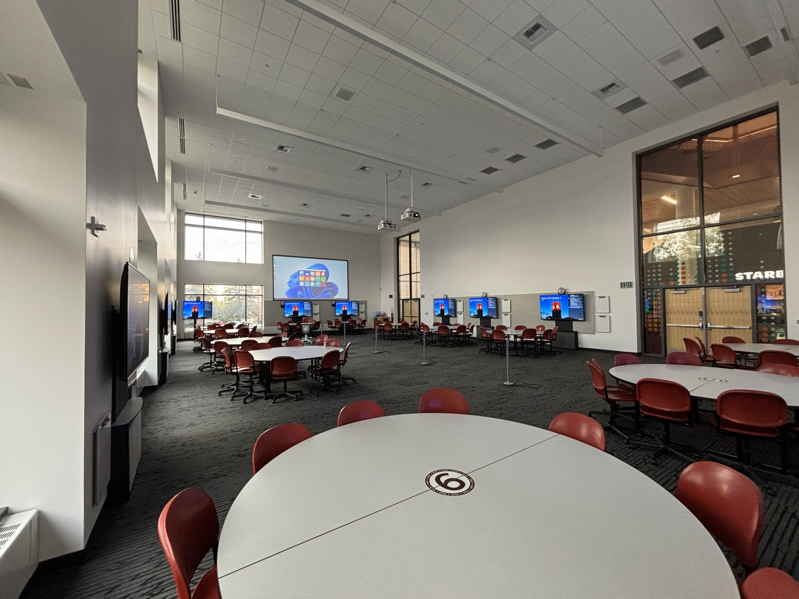 Looking out from the corner of the classroom over round tables and moveable student seating and toward the side of the room where the instructor teaching station and one large projection screen are located. Each of the twelve student groups consists of a round table with nine chairs and has a large monitor for viewing content. Two sets of double doors can be seen in the right front and back corners of the room and there are several convenience power towers available for pugging in devices in the middle of the room.