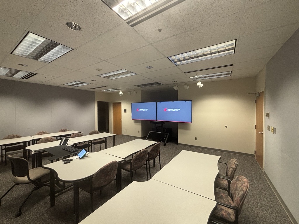 View from the back of the classroom showing four rows of tables with chairs facing the center of the room. In the middle of the tables is an instructor seat with access to technology. At the front of the room, two large monitors are mounted on a stand next to a door. Another door is located on the right side wall.