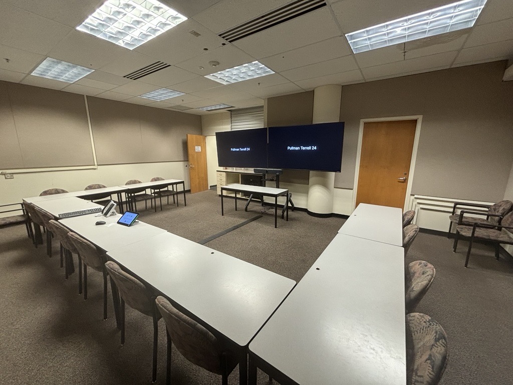 View from the back of the classroom showing seven tables arranged in a U‑shape, each with two chairs facing the front and center of the room. In the center of the U‑shaped layout is a table for the instructor equipped with technology. At the front of the room, two large monitors are mounted on a stand with a door on either side.