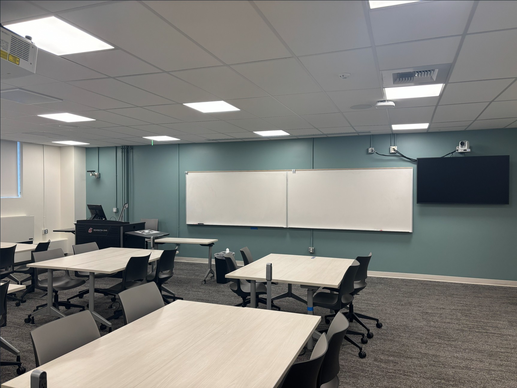 View from the back corner of the classroom showing five groups of movable tables and chairs. At the front of the room, an instructor podium with technology sits next to a large white board and a wall mounted monitor with a camera above it.