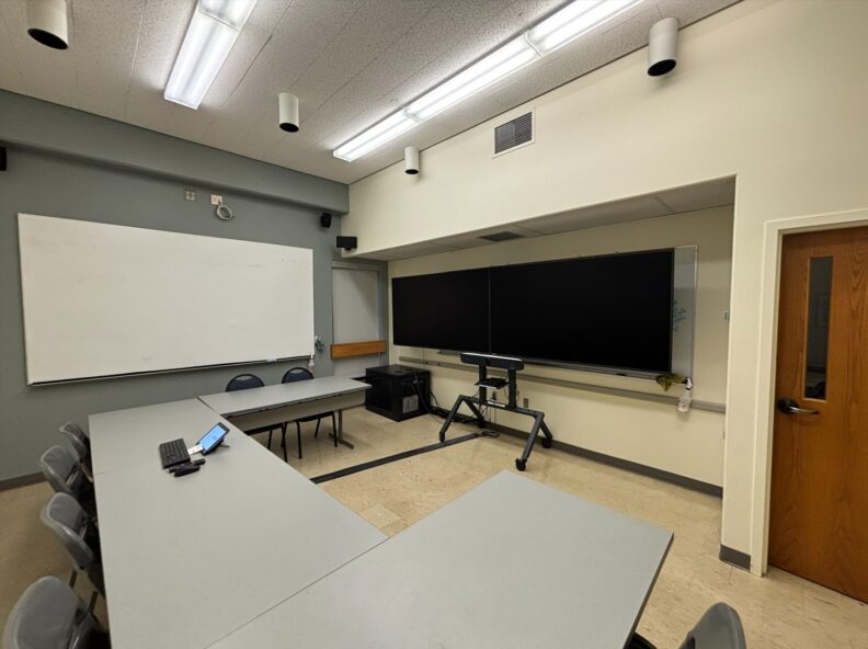Classroom viewed from the back with four rectangular tables arranged in a U‑shape. Instructor equipment sits on the center table. Chairs face the front of the room, which has two monitors on a stand with a camera below. A whiteboard is mounted on the left wall, and a door is to the right of the monitors.