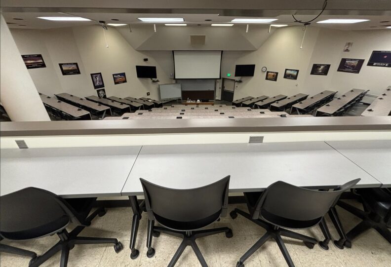 View from the back row of the large, tiered lecture hall with long tables that include power outlets and rolling chairs. At the front of the room are a projection screen, two large monitors with cameras mounted below them, a wall-mounted whiteboard, and a rolling whiteboard. A ground‑level entry door is on the right side of the room.