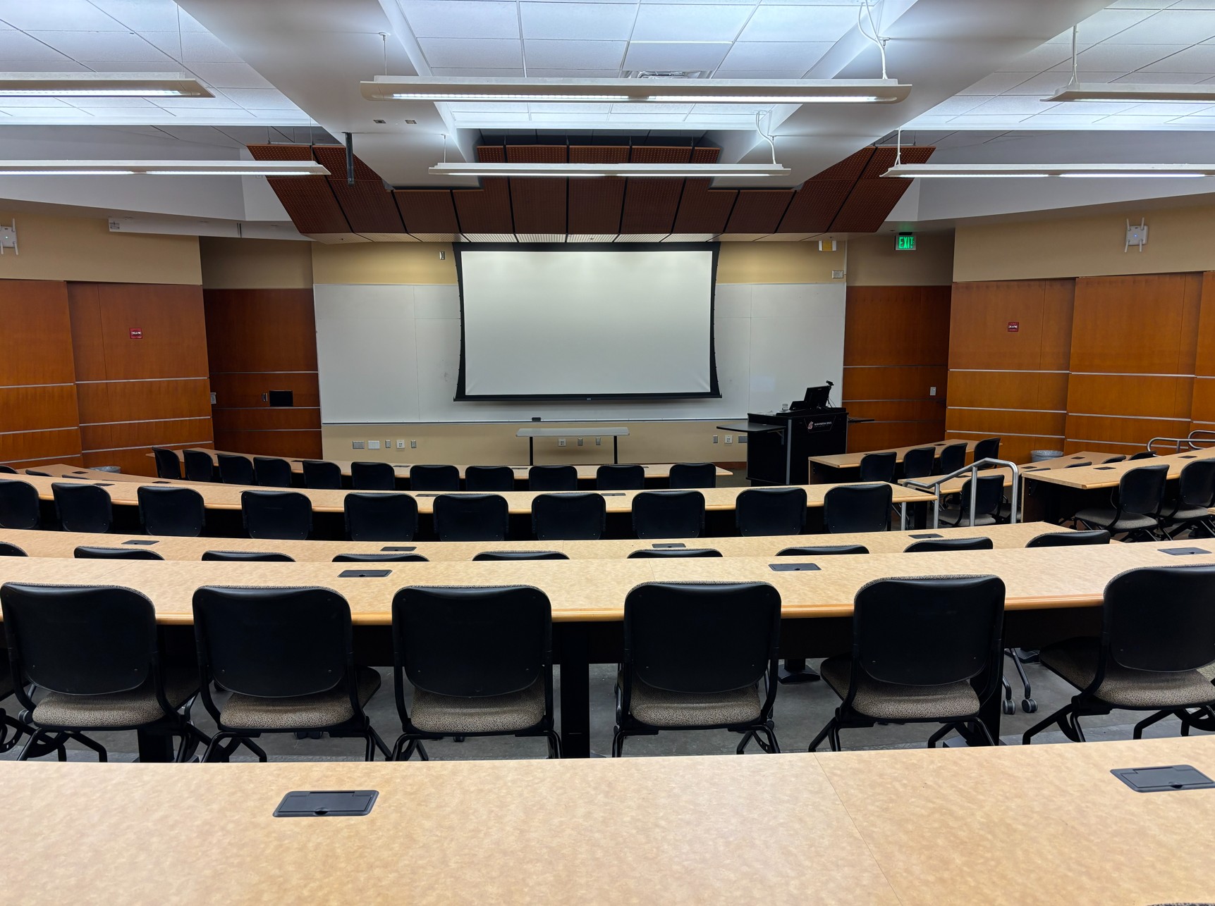 Looking out from the rear of the classroom over the rows of student seating and toward the front of the room where the instructor's podium, one whiteboard, and one projection screen are located.