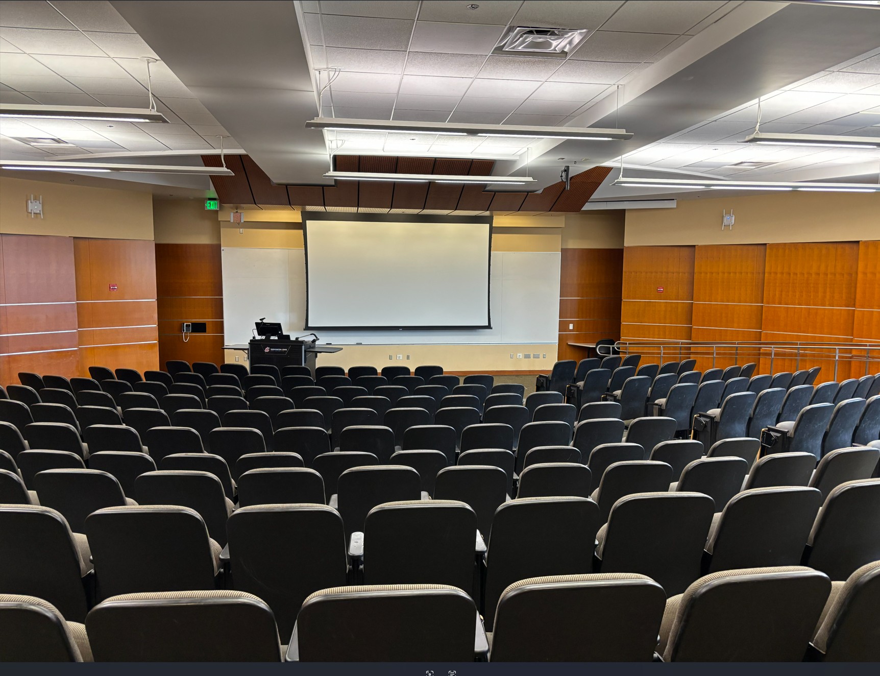 Looking out from the rear of the classroom over the rows of student seating and toward the front of the room where the instructor podium, one whiteboard, and one projection screen are located.