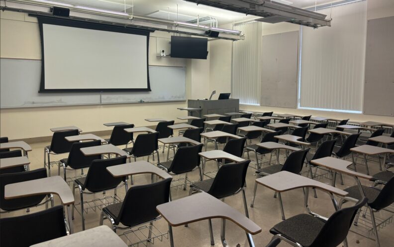 View from the rear of the classroom looking over rows of movable student seating. At the front of the room are the instructor’s podium with integrated technology, a projection screen, a large monitor with a camera mounted below it, and a wall‑mounted whiteboard that spans the front wall.