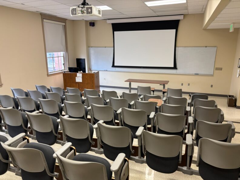 View from the back of the classroom over the rows of fixed student seating with foldable tablet arms facing forward. At the front of the room are a wall mounted whiteboard, a projection screen, and an instructor lectern with integrated technology.