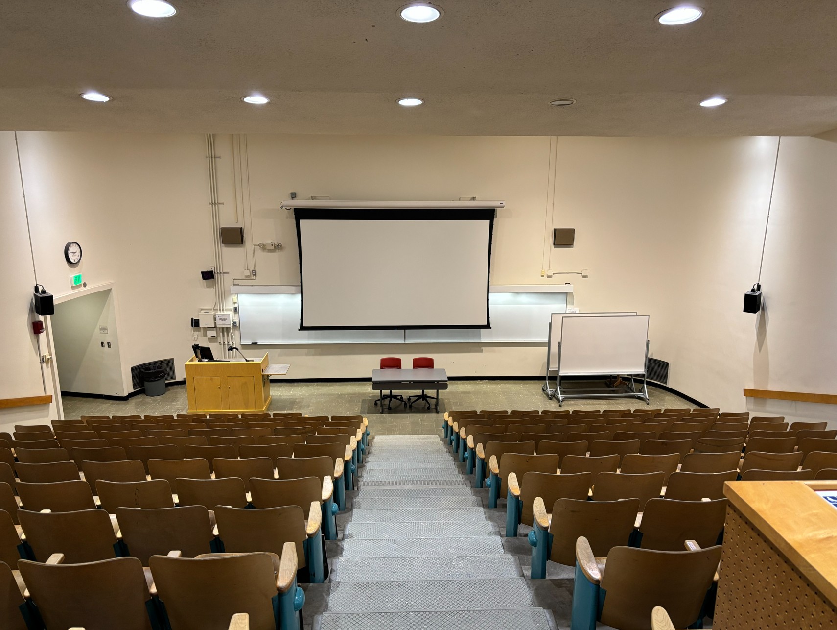 Looking out from the rear of the classroom over the rows of student seating and toward the front of the room where the instructor's podium, one display screen, one wall-mounted whiteboard, and two rolling whiteboards are located. Sets of stairs flank the left and right bank of seats, and a center aisle divides the seating into two banks.
