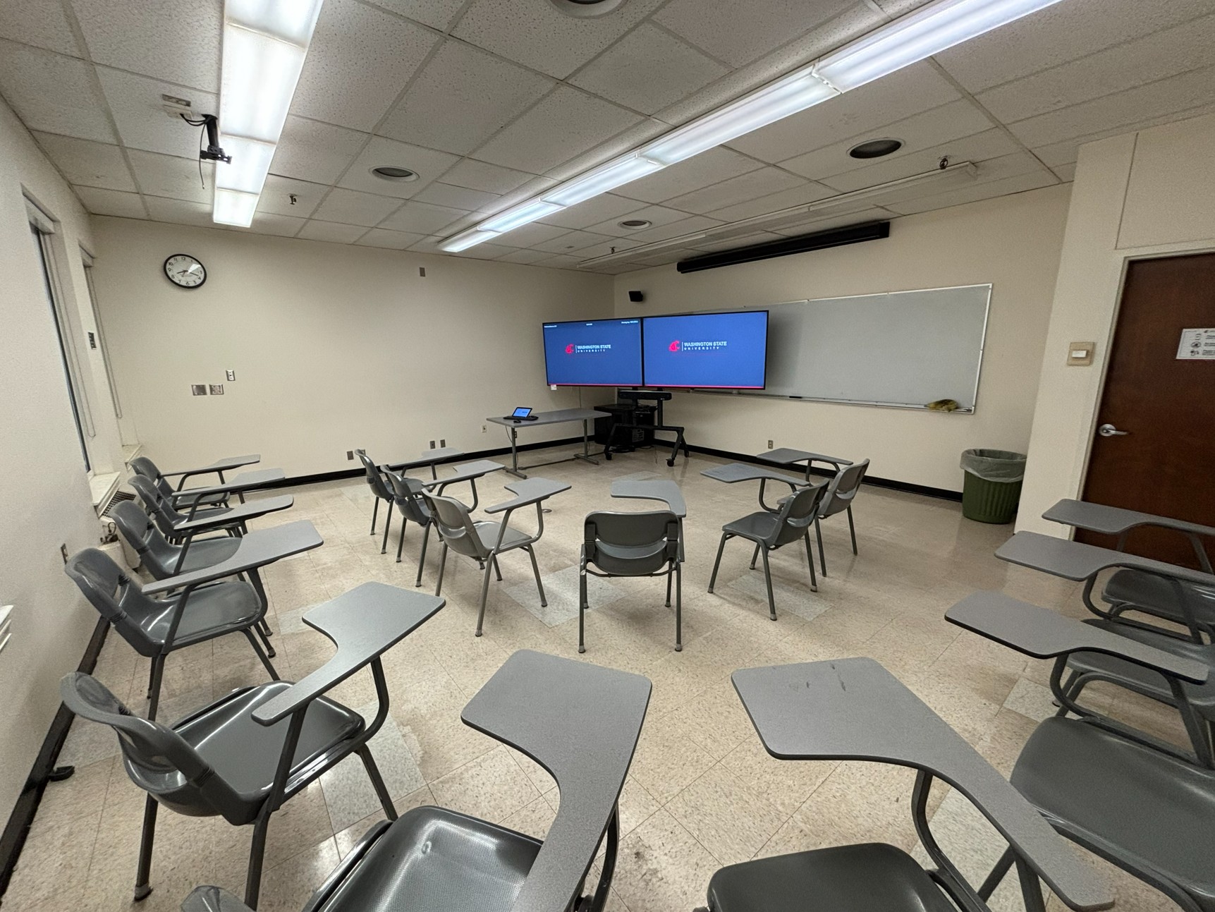 View from back of classroom show two u-shaped rows of moveable student seating. At the front there is a wall-mounted whiteboard, table with instructor technology, and two monitors on a stand with a camera below it.