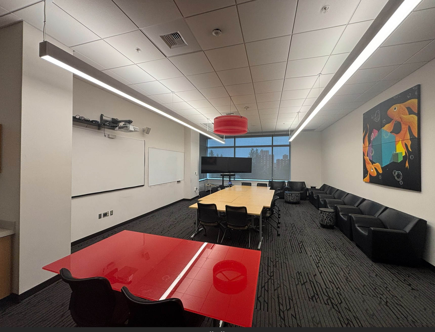 View from left of room showing two tables and various movable seating options. At the front is a projection screen, a wall mounted whiteboard, and two large monitors on a movable stand with cameras beneath them. The room features student artwork, including a red chandelier and a large abstract painting on the back wall.