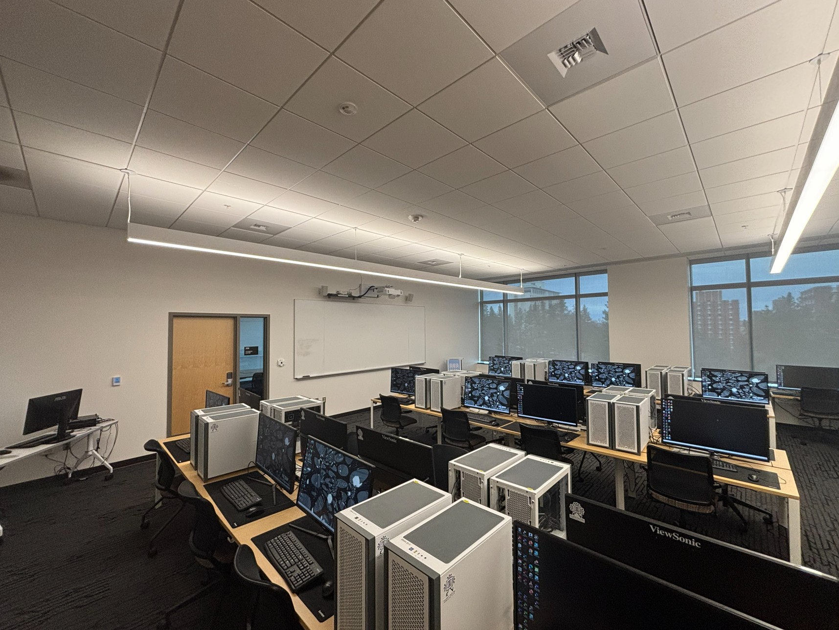 View from the back of a computer‑equipped classroom showing three rows of workstations, each with eight computers where students sit facing one another. At the front is an instructor station with integrated technology and a projection screen that doubles as a whiteboard. A door to an adjoining room is located between these features.
