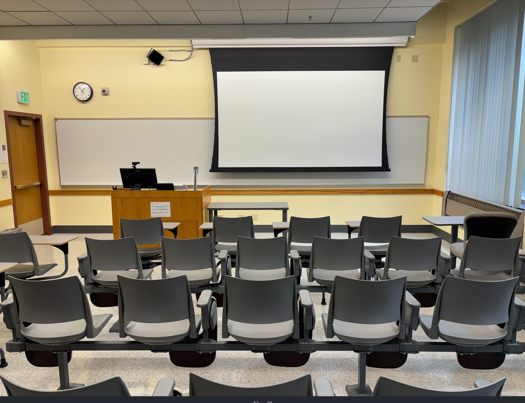 View from the rear of the room looking over four rows of movable student seating. At the front of the room are an instructor table and podium with integrated technology, a projection screen, and a whiteboard that spans the front wall. A door is located on the left side of the room.