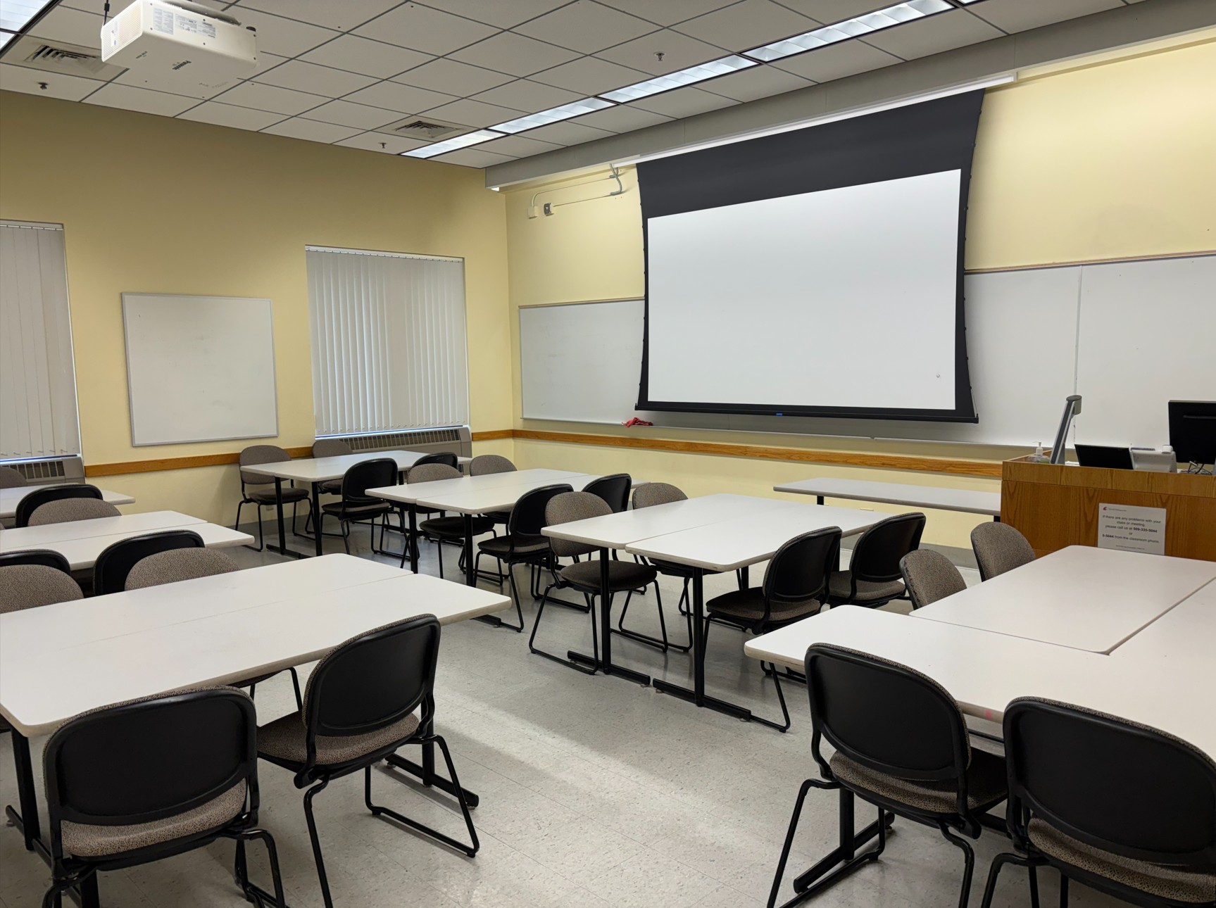 View from the back corner of the classroom showing seven groups of tables with movable chairs around them. At the front of the room are a wall‑mounted whiteboard, a projection screen, and an instructor lectern with integrated technology. A smaller student whiteboard is mounted on the left wall.