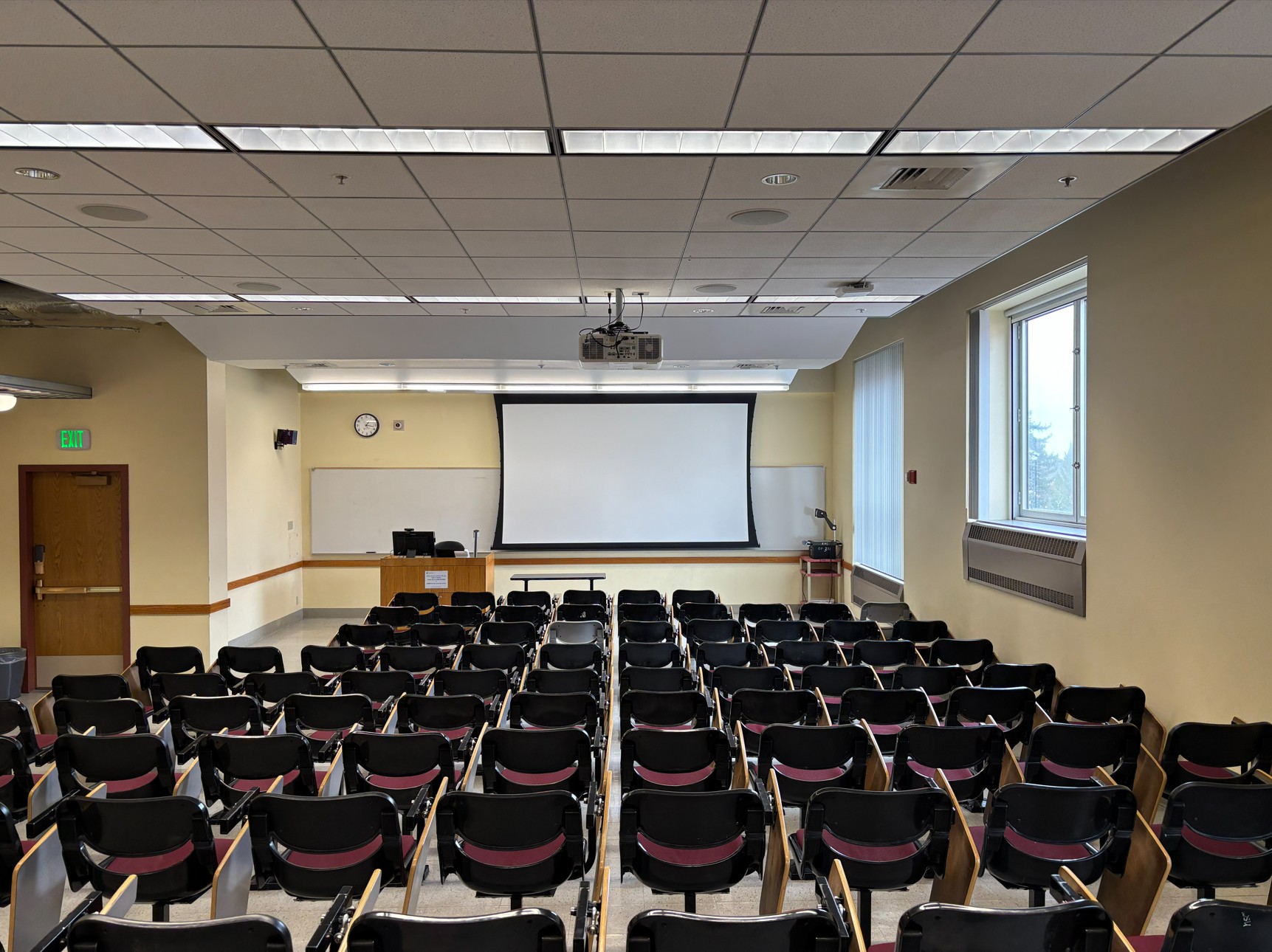 View from the back of the classroom showing rows of fixed student seating facing the front of the room where an instructor podium with integrated technology, projection screen, overhead projector, and wall-mounted whiteboard are located. There is a door to the left side of the room.