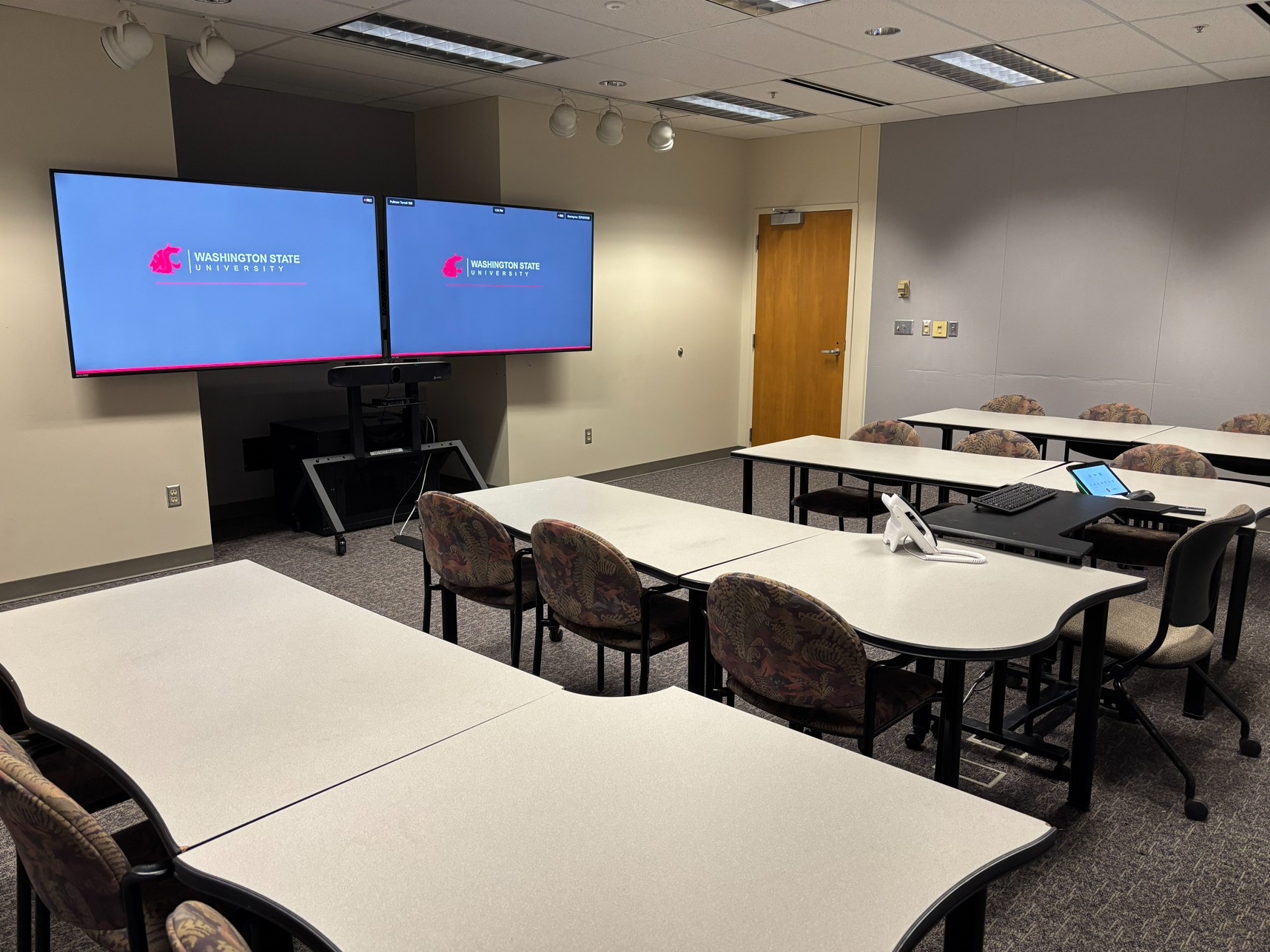 View from the back of classroom showing four rows of tables with moveable chairs facing center. In the middle of the tables is an instructor seat with access to technology. At the front, two large monitors are mounted on a stand with cameras below. A door is located on the right wall.