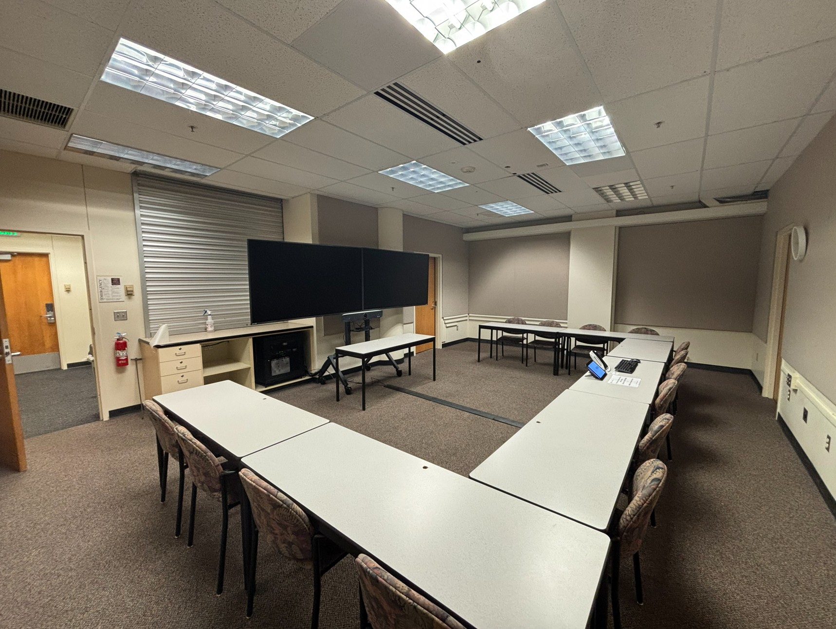 View from the back of the classroom showing seven tables arranged in a U‑shape, each with two chairs facing the front or center of the room. In the center of the U‑shaped layout is a table for the instructor equipped with technology. At the front of the room, is table in front of two large monitors mounted on a moveable stand with cameras below. There are three doors, two at the front and one at the back.