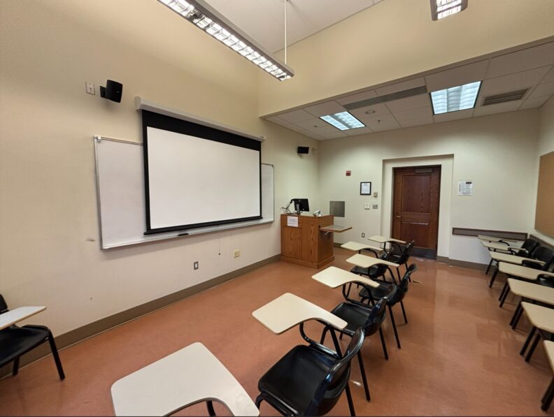 View from the rear of the room looking over two rows of movable student seating. At the front of the room are an instructor podium with integrated technology, a projection screen, and a whiteboard that spans the front wall. A door is located on the right side of the room.