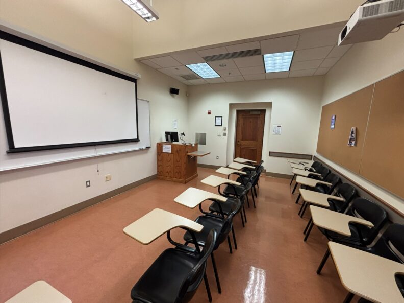 View from the rear of the room looking over two rows of movable student seating. At the front of the room are an instructor podium with integrated technology, a projection screen, and a whiteboard that spans the front wall. A door is located on the right side of the room.