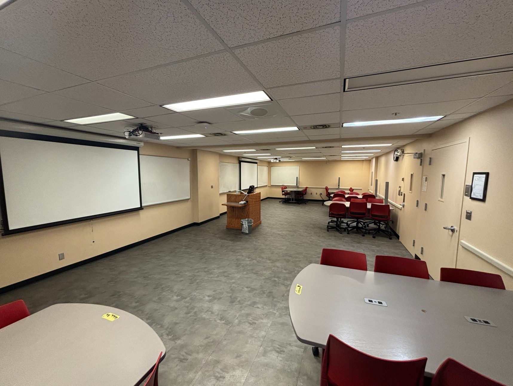 View from a corner of the classroom showing seven tables, each with six movable chairs. At the front of the room is an instructor lectern equipped with technology and two projection screens, one on each side.