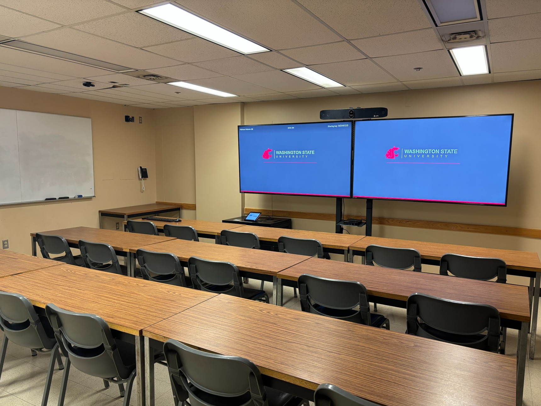 View from the back of the classroom showing three rows of movable tables and chairs facing the front. The far-left seat in the first row includes instructor technology. At the front of the room are two large monitors on a stand with a camera mounted above them. Wall-mounted whiteboards line the left side wall.