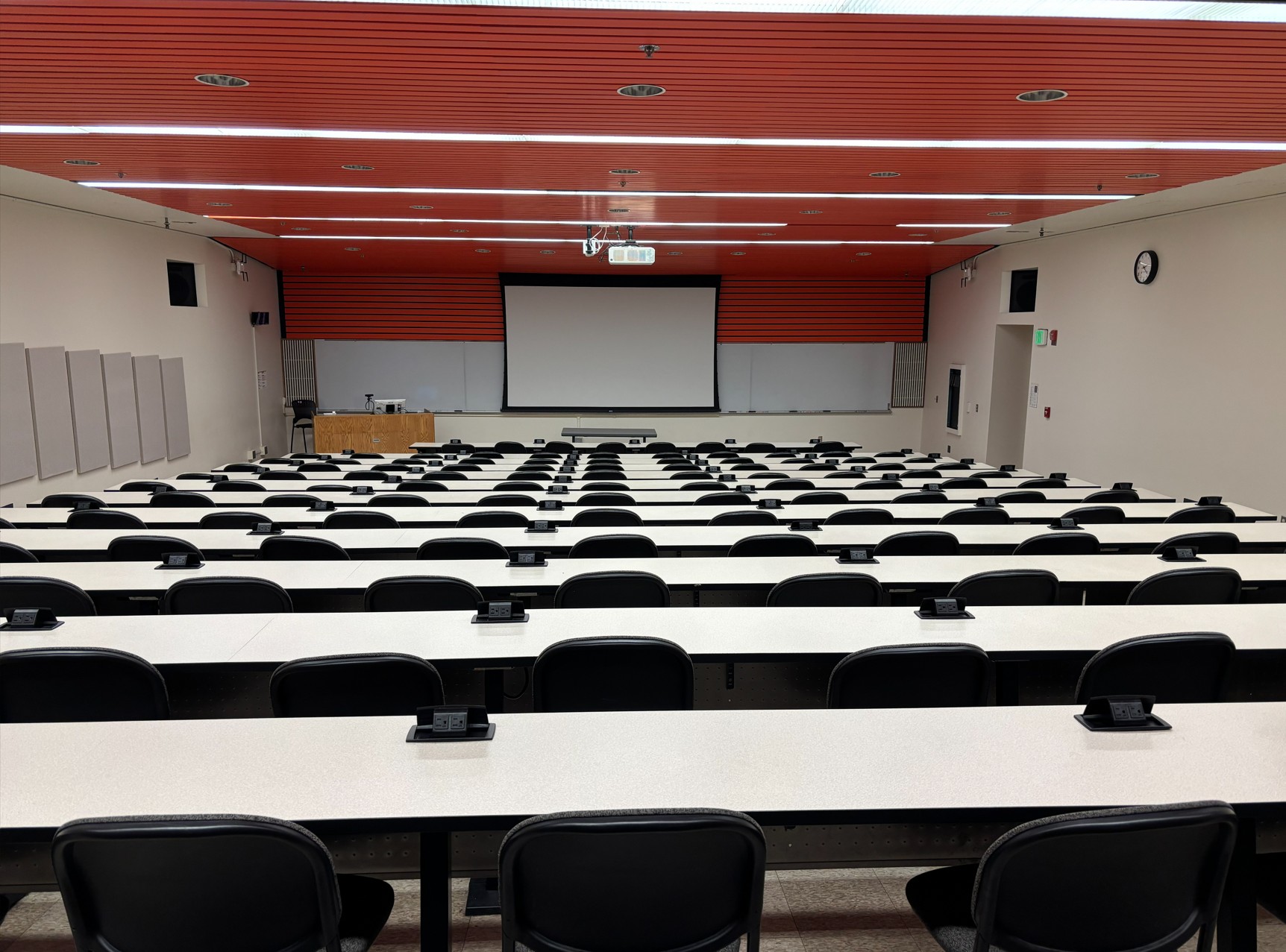 View from the back of a sloped lecture hall showing rows of fixed tables with power outlets and swivel chairs. At the front of the room are a wall mounted whiteboard, a projection screen, and an instructor lectern with integrated technology. A ground level exit door is located on the right wall.
