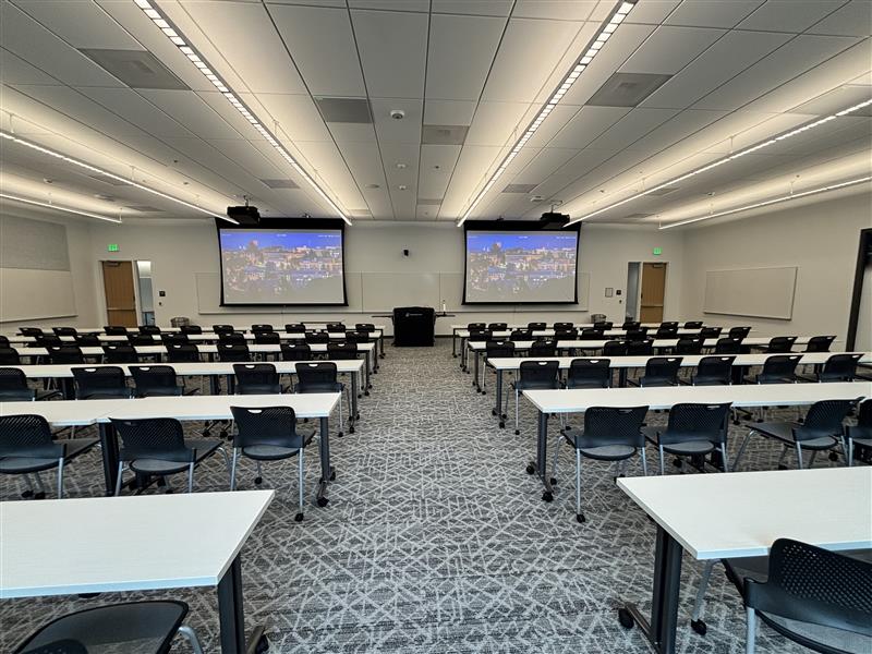 Classroom viewed from the back with six rows of movable tables and chairs divided by a center aisle facing the front. An instructor podium with technology controls sits at the front between two projection screens. Whiteboards cover the front and side walls, with a camera mounted above the front boards. Exit doors are on both sides of the front wall.