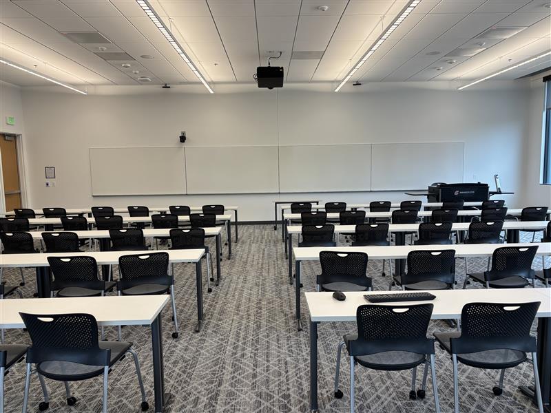 Classroom viewed from the back with five rows of movable tables and chairs divided by a center aisle facing the front. An instructor podium with technology controls sits at the front. Whiteboards cover the front wall with a camera mounted above them, and an exit door is at the front left.