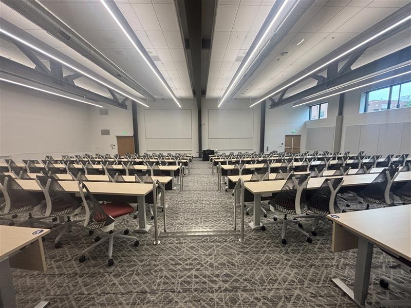 Tiered classroom viewed from the back with rows of fixed tables and movable chairs divided by a center aisle facing the front. An instructor podium with technology controls sits between two projection screens. Whiteboards span the front wall, with exit doors on either side.