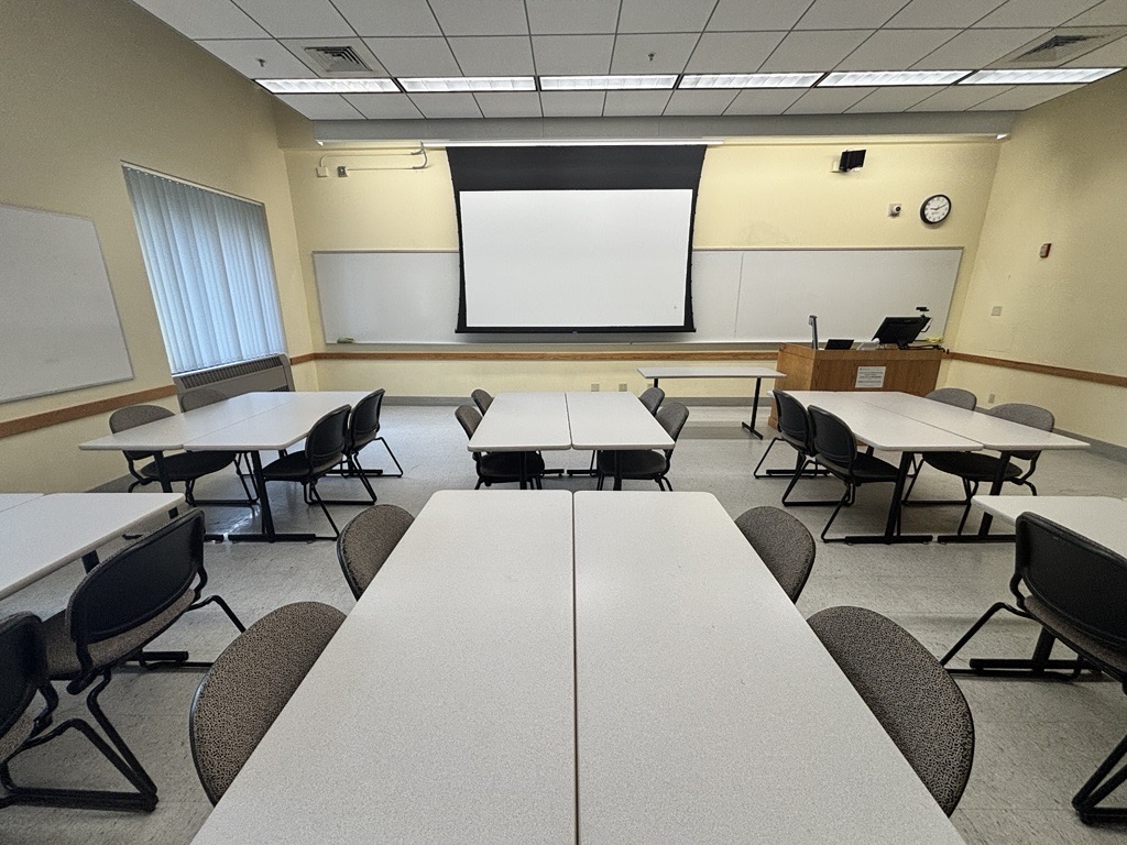Classroom viewed from the back, showing six groups of tables and chairs arranged for face-to-face discussion. At the front right are an instructor podium, classroom technology, and a projection screen. Whiteboards line the front and side walls.