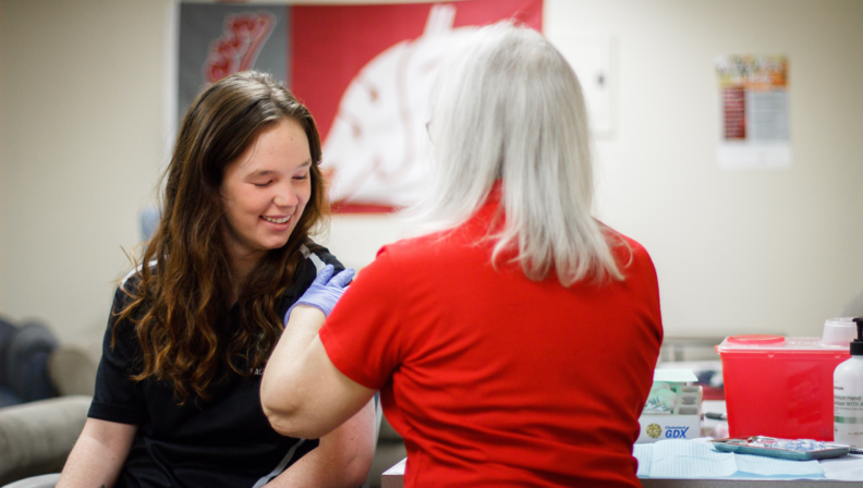 A young woman receiving a vaccination from a healthcare worker wearing gloves inside a clinic setting. A Washington State University flag hangs in the background.