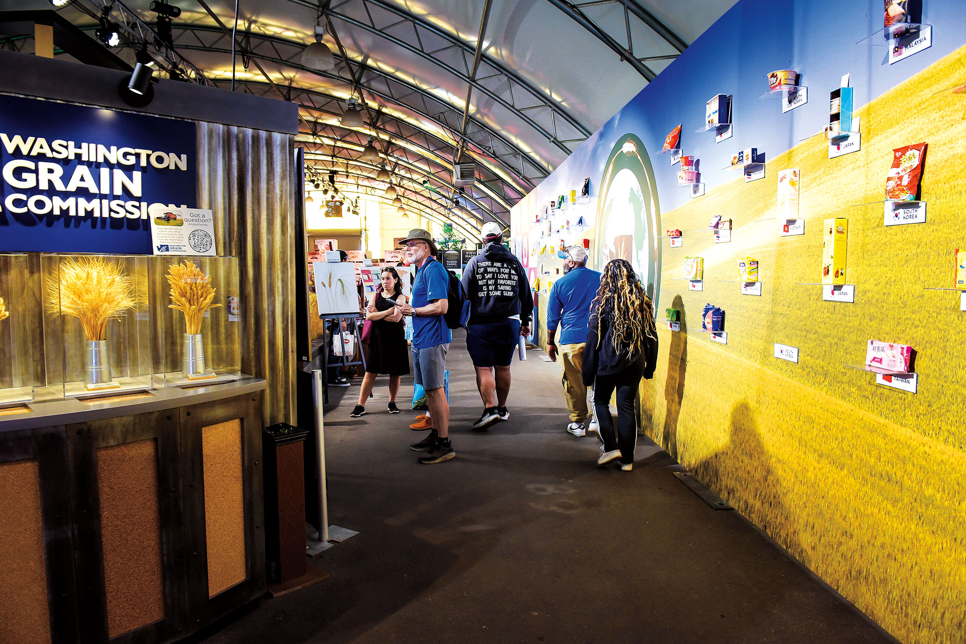 Individuals are walking through a display pavilion with information about Washington agriculture. This image features a display from the Washington Grain Commission with stalks of wheat.