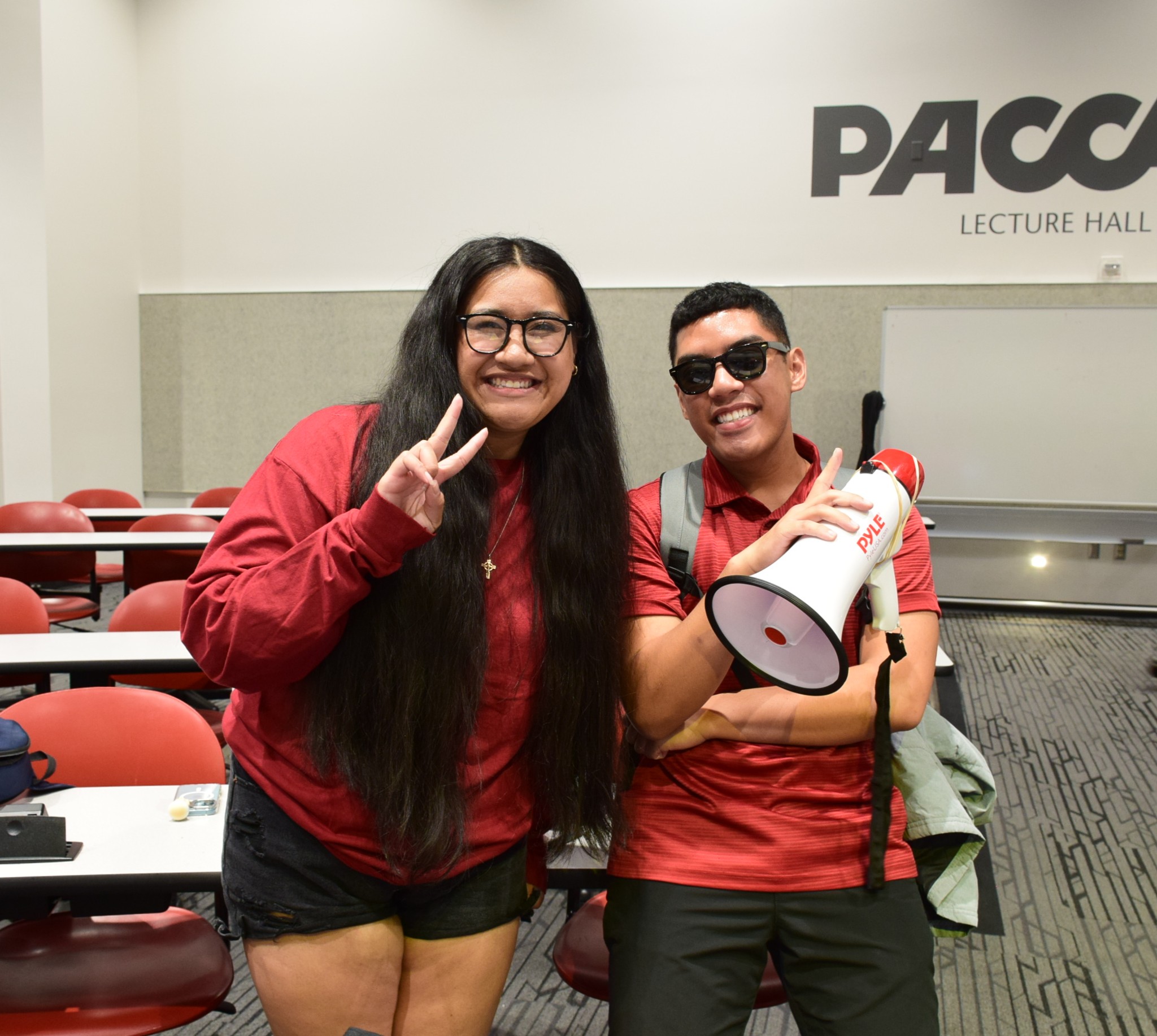 A female and male student are in a lecture hall, smiling and facing the camera. One student has a megaphone and is wearing sunglasses.
