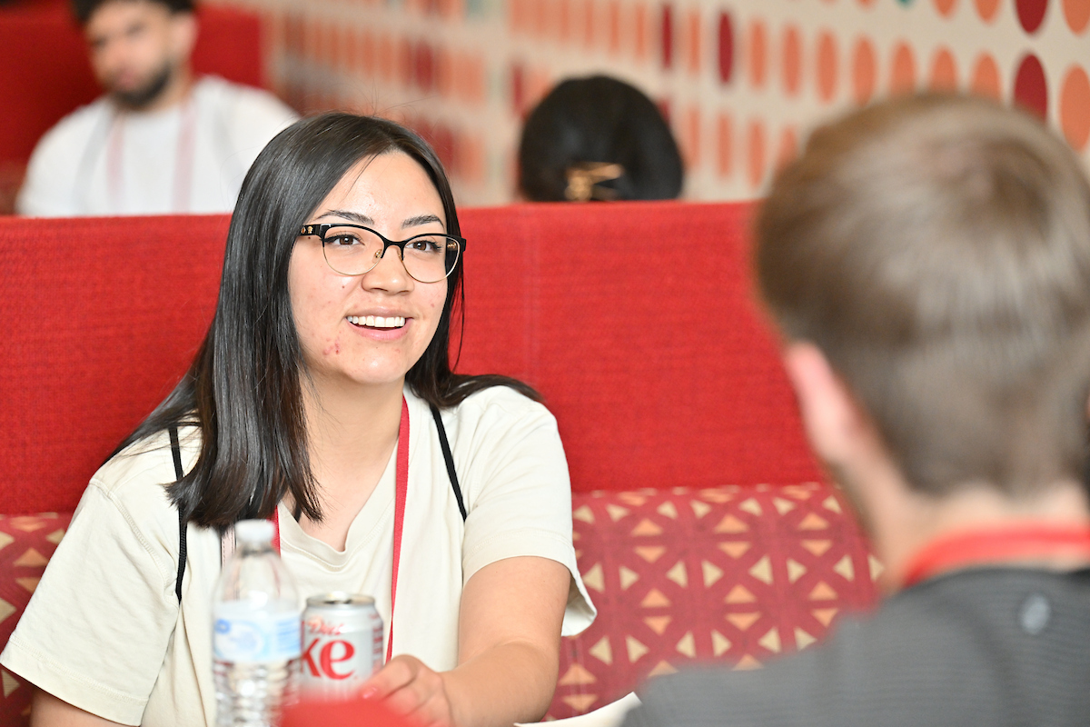 A female student with glasses sitting in a red seating booth talking with someone who is facing her in the foreground.